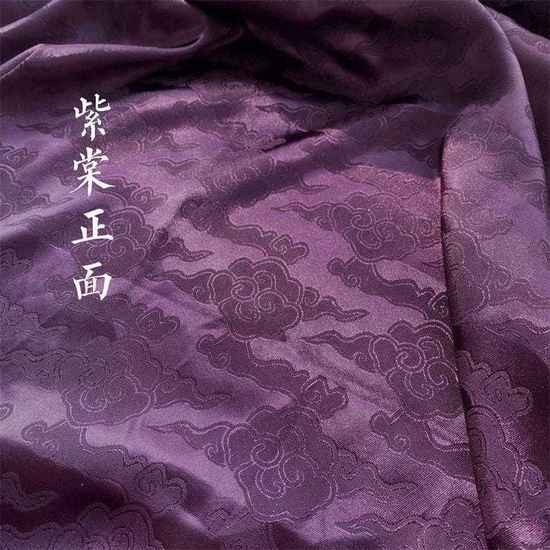 May include: A deep purple fabric featuring a tonal jacquard pattern of stylized clouds. The material has a smooth, lustrous finish with visible folds and drapes. The Chinese characters 紫棠正面 are printed vertically in white on the left side.