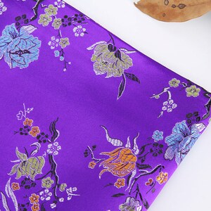 May include: Purple fabric with a floral pattern. The flowers are in shades of orange, green, blue, and white. The fabric has a satin-like sheen.