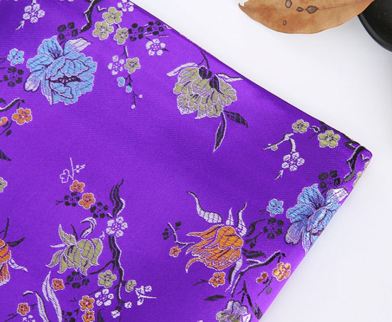 May include: Purple fabric with a floral pattern. The flowers are in shades of orange, green, blue, and white. The fabric has a satin-like sheen.