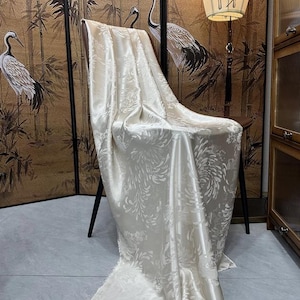 May include: A white satin fabric with a floral pattern draped over a chair. The fabric has a soft, luxurious feel and is perfect for upholstery or drapery.