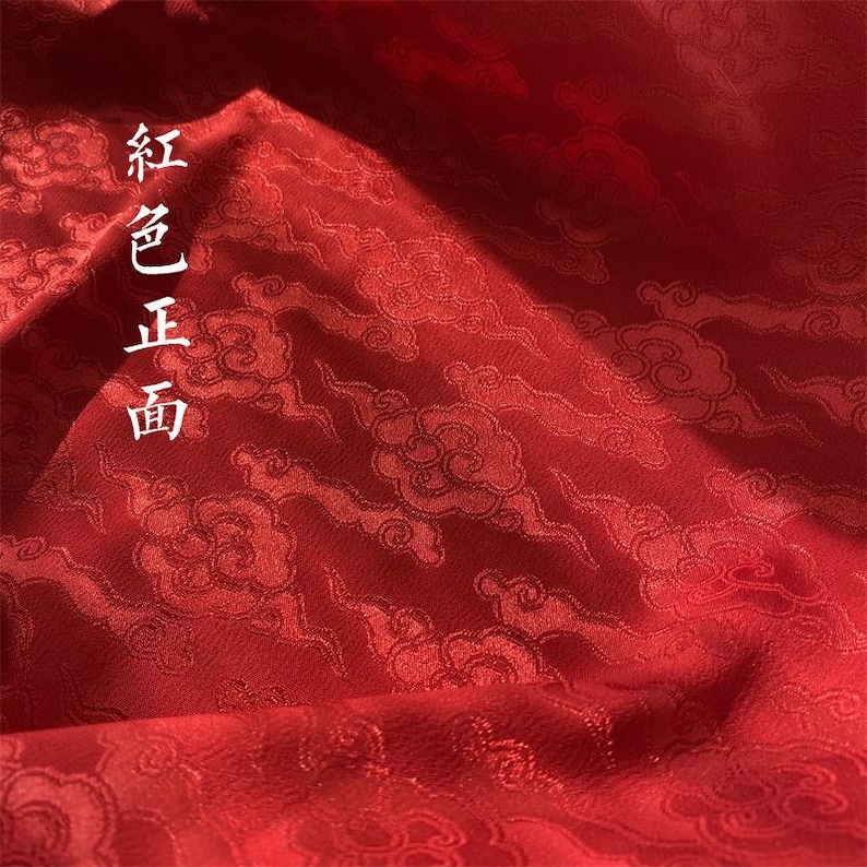 May include: A deep red fabric featuring a repeating woven pattern of stylized clouds. The surface has a subtle sheen and includes the vertical Chinese characters 紅色正面 printed in white on the left side of the textile material.