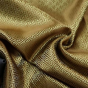 Könnte beinhalten: Nahaufnahme eines braunen Stoffes mit einem goldenen geometrischen Muster. Der Stoff ist gefaltet und über eine Holzoberfläche gelegt.