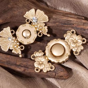 May include: Two gold-colored decorative clasps with floral and fan-shaped designs. Each clasp features a central circular element and is adorned with small crystals. The clasps are set against a textured wooden surface and a gold fabric.