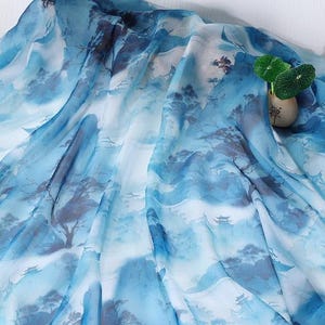 May include: A flowing fabric with a watercolor-style landscape design in shades of blue and white. The pattern depicts mountains, trees, and small structures, creating a serene and artistic aesthetic. A small vase with green leaves is on the right side.