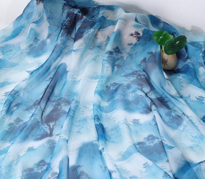 May include: A flowing fabric with a watercolor-style landscape design in shades of blue and white. The pattern depicts mountains, trees, and small structures, creating a serene and artistic aesthetic. A small vase with green leaves is on the right side.