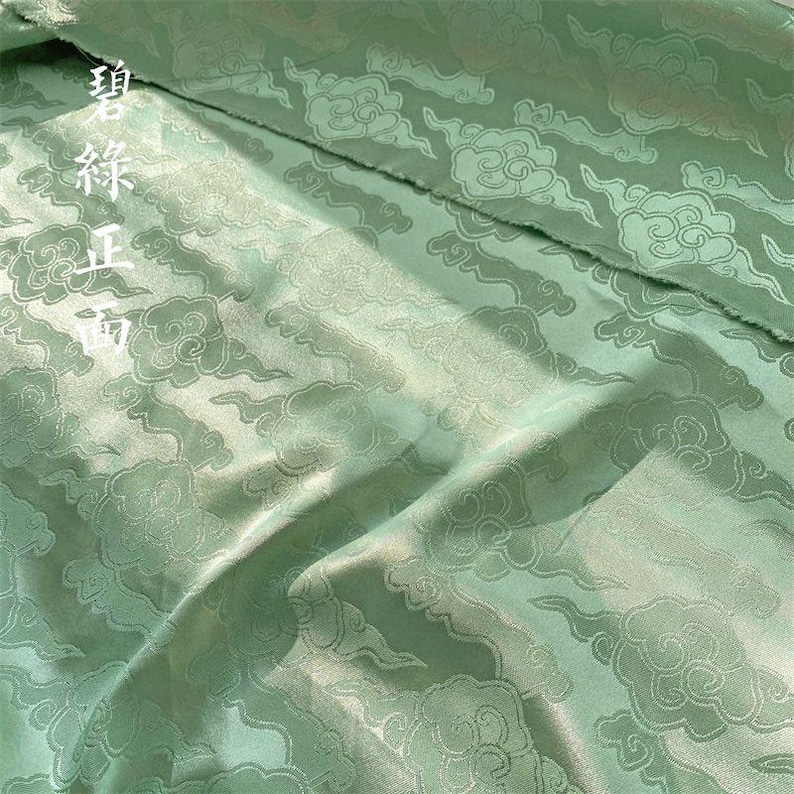 May include: A light green fabric featuring a woven pattern of stylized clouds. The material has a subtle sheen and includes white Chinese characters printed on the upper left side, which read 碧綠正面, indicating the front side of the green fabric.