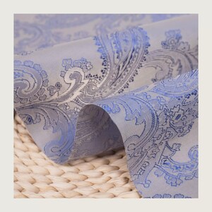 May include: Close-up of a folded fabric with a paisley pattern. The fabric is a light gray color with a blue paisley design. The fabric is soft and has a slight sheen. The background is a woven texture.
