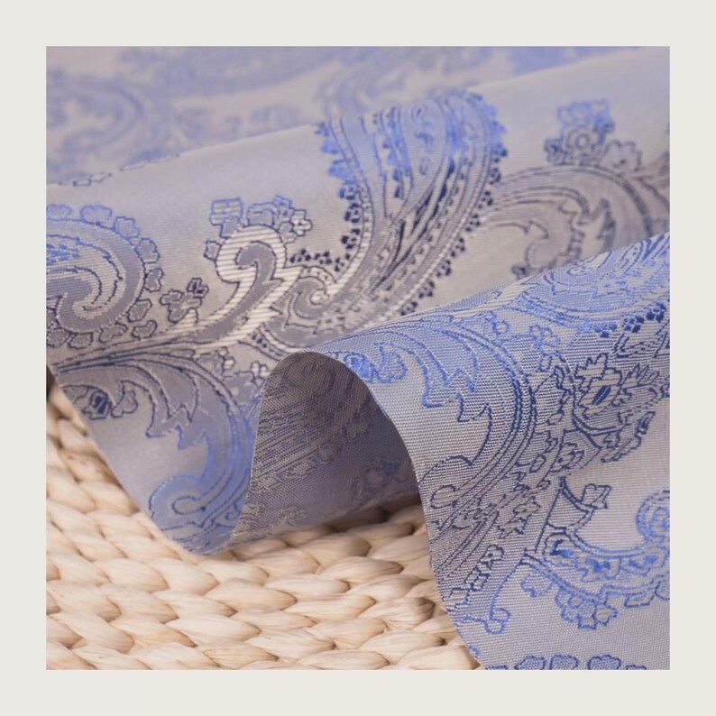 May include: Close-up of a folded fabric with a paisley pattern. The fabric is a light gray color with a blue paisley design. The fabric is soft and has a slight sheen. The background is a woven texture.