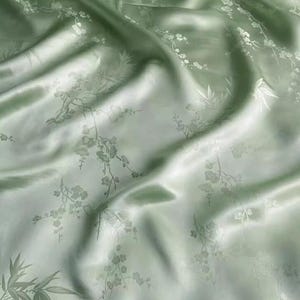 Puede incluir: Una tela de color verde salvia claro con un acabado satinado y brillante. El material presenta un patrón tejido tonal que representa delicadas ramas de cerezo y hojas de bambú, creando una apariencia sutil y texturizada sobre la superficie drapeada.