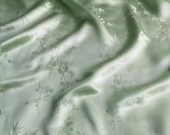 May include: A light sage green fabric with a lustrous, satin-like finish. The material features a tonal, woven pattern depicting delicate cherry blossom branches and bamboo leaves, creating a subtle, textured appearance across the draped surface.
