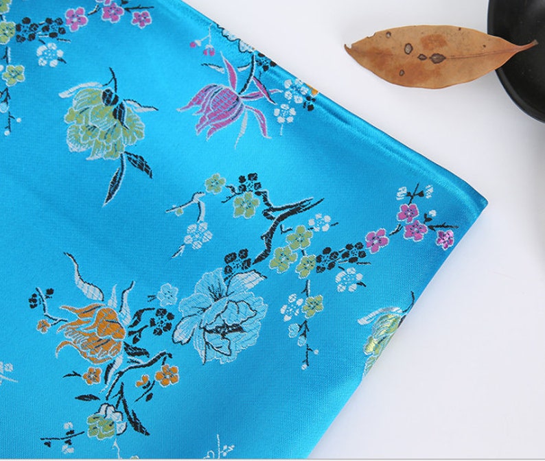 May include: A blue fabric with a floral pattern. The fabric has a satin finish and features a variety of flowers in shades of pink, yellow, and green.
