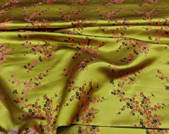 Op de afbeelding: Een lengte glanzende chartreuse stof met een herhalend patroon van kleine, delicate kersenbloesemtakken in koraalroze en zwarte tinten. Het materiaal heeft een gladde, satijnachtige afwerking met zichtbare plooien en draperingen.