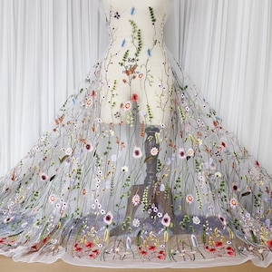 May include: White sheer fabric with embroidered floral designs in various colors, including red, pink, blue, and green. The fabric is displayed on a dress form.