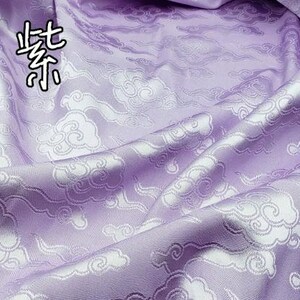 May include: A light purple fabric featuring a repeating woven pattern of white stylized clouds. The Chinese characters 浅紫 are printed in the upper left corner of the textile, which is draped to show folds and a smooth, slightly reflective surface texture.
