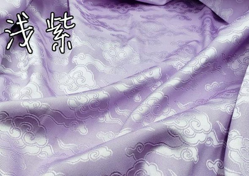 May include: A light purple fabric featuring a repeating woven pattern of white stylized clouds. The Chinese characters 浅紫 are printed in the upper left corner of the textile, which is draped to show folds and a smooth, slightly reflective surface texture.
