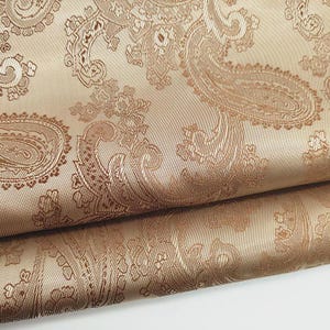May include: Close-up of a folded fabric with a paisley pattern. The fabric is a light tan color with a darker brown paisley design. The paisley pattern is detailed and intricate, with swirling shapes and floral elements.