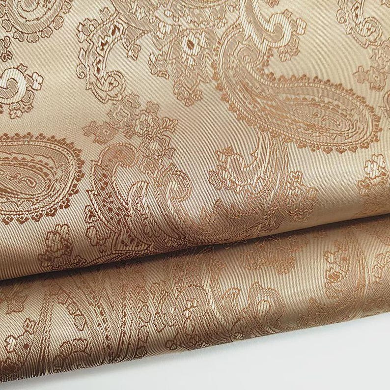 May include: Close-up of a folded fabric with a paisley pattern. The fabric is a light tan color with a darker brown paisley design. The paisley pattern is detailed and intricate, with swirling shapes and floral elements.