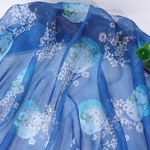 May include: A sheer, flowing fabric in shades of blue, adorned with a repeating pattern of light blue circles, white floral branches, and small birds. The fabric is lightweight and appears to be silk or a similar material.