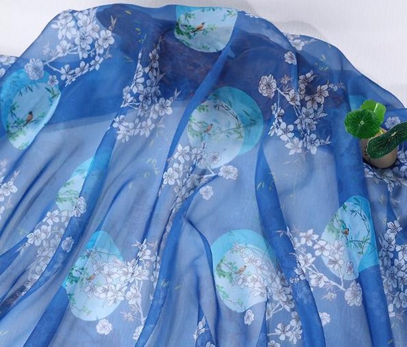 May include: A sheer, flowing fabric in shades of blue, adorned with a repeating pattern of light blue circles, white floral branches, and small birds. The fabric is lightweight and appears to be silk or a similar material.