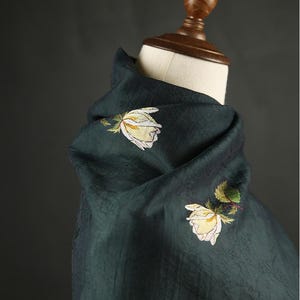 Pu&ograve; includere: Sciarpa verde scuro con rose bianche e gialle ricamate. La sciarpa &egrave; drappeggiata su una testa di manichino, mettendo in risalto la consistenza del tessuto e il design floreale. Il colore della sciarpa &egrave; un verde intenso e ricco.