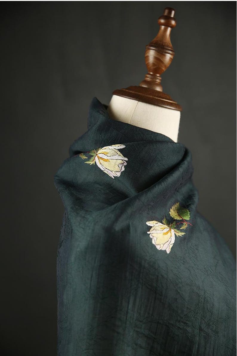 Pu&ograve; includere: Sciarpa verde scuro con rose bianche e gialle ricamate. La sciarpa &egrave; drappeggiata su una testa di manichino, mettendo in risalto la consistenza del tessuto e il design floreale. Il colore della sciarpa &egrave; un verde intenso e ricco.