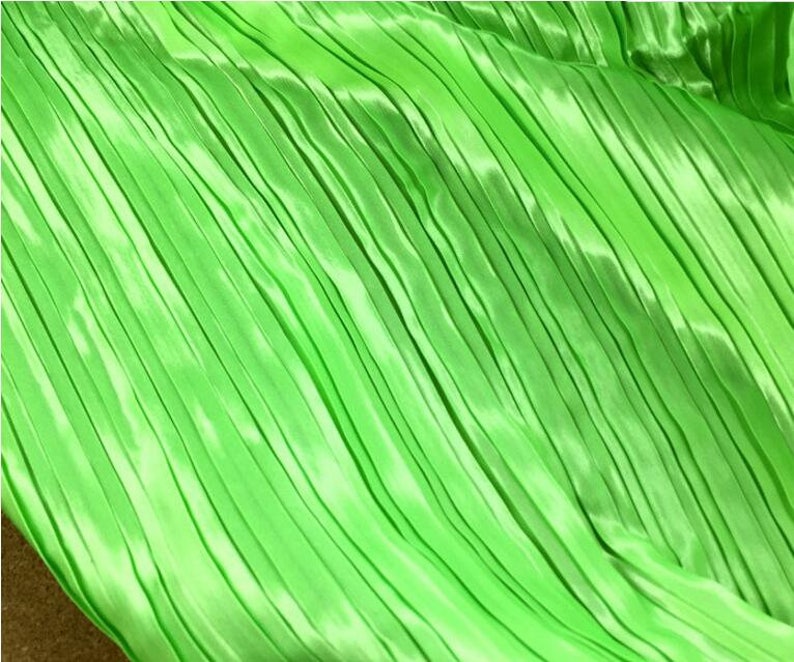 May include: A close-up of a bright green pleated fabric. The fabric has a smooth, silky texture and is folded in a diagonal pattern.