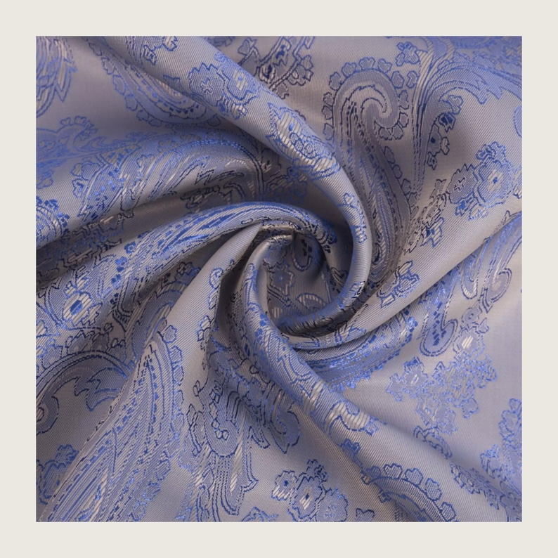 May include: Close-up of a folded fabric with a paisley pattern. The fabric is a light gray color with a blue paisley design. The fabric is soft and has a slight sheen.