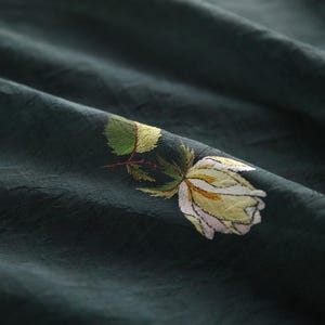 Pu&ograve; includere: Tessuto verde scuro con una rosa ricamata bianca e gialla. La rosa ha foglie verdi e uno stelo rosso. Il tessuto ha un aspetto strutturato con pieghe sottili, creando un senso di profondit&agrave; e movimento.