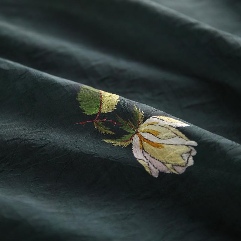 Pu&ograve; includere: Tessuto verde scuro con una rosa ricamata bianca e gialla. La rosa ha foglie verdi e uno stelo rosso. Il tessuto ha un aspetto strutturato con pieghe sottili, creando un senso di profondit&agrave; e movimento.