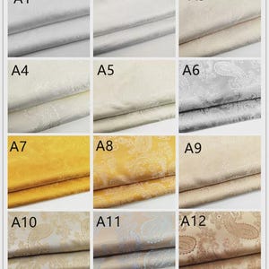 May include: A collection of fabric swatches in various colors, including silver, cream, gold, and beige. Each swatch displays a paisley pattern. The fabrics are neatly folded, showcasing their texture and design. The image is well-lit, highlighting the different shades and patterns.