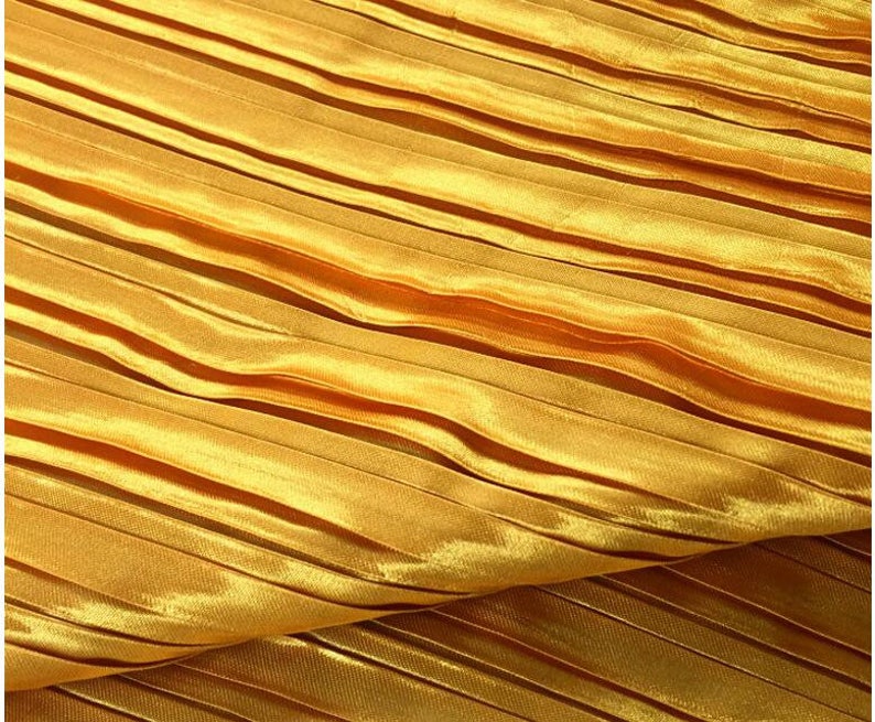 May include: A close-up of a gold pleated fabric. The fabric has a smooth, shiny surface and is folded in a wavy pattern.