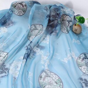 May include: A light blue silk scarf with a watercolor design. The scarf features a landscape pattern with mountains, trees, and geometric shapes. White floral accents are scattered across the fabric. A small decorative vase with a plant is in the background.