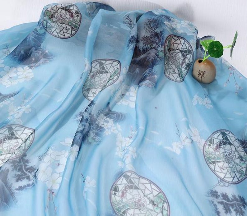 May include: A light blue silk scarf with a watercolor design. The scarf features a landscape pattern with mountains, trees, and geometric shapes. White floral accents are scattered across the fabric. A small decorative vase with a plant is in the background.
