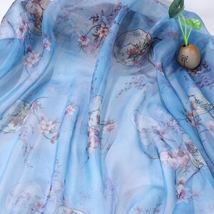 May include: A flowing, light blue fabric with a delicate floral pattern in shades of pink and green. The fabric has a semi-transparent quality, with a subtle sheen. The design includes oriental-style illustrations and is suitable for scarves or apparel.