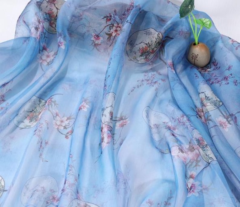 May include: A flowing, light blue fabric with a delicate floral pattern in shades of pink and green. The fabric has a semi-transparent quality, with a subtle sheen. The design includes oriental-style illustrations and is suitable for scarves or apparel.
