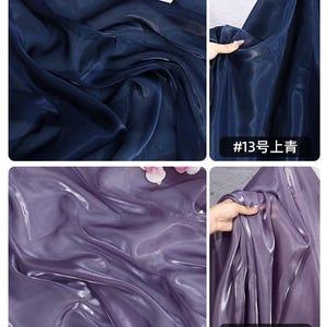 May include: Close-up of shimmering fabric in two colors: deep navy blue and a soft lavender-purple. The fabric has a flowing, translucent quality, with light reflecting off its surface. The image includes text in the bottom right corner.