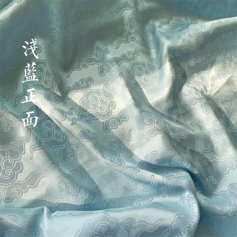 May include: A light blue fabric featuring a repeating woven pattern of stylized clouds. The material has a smooth, satin-like sheen with visible folds and drapes. The Chinese characters 浅藍正面 are printed vertically in white on the left side.