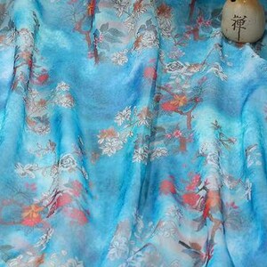 May include: A flowing, light blue fabric with a floral pattern in white, orange, and red. The fabric has a watercolor effect, with soft folds and a subtle sheen. A small, beige, cylindrical object with Japanese characters is on the fabric.