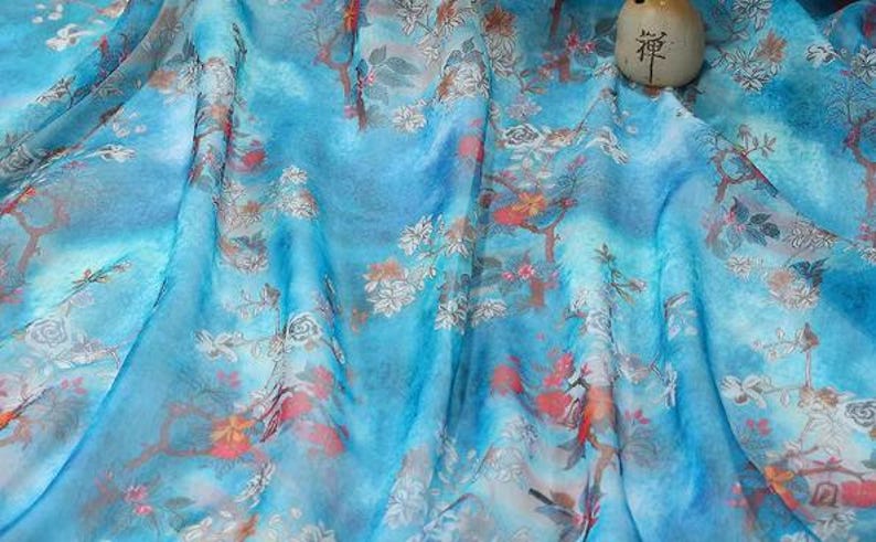 May include: A flowing, light blue fabric with a floral pattern in white, orange, and red. The fabric has a watercolor effect, with soft folds and a subtle sheen. A small, beige, cylindrical object with Japanese characters is on the fabric.