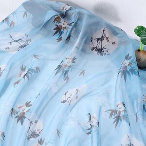 May include: Light blue sheer fabric with a floral and landscape print. The fabric features white circular designs with black bamboo and floral accents in brown and white. The fabric is draped, creating soft folds.