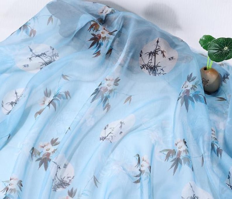 May include: Light blue sheer fabric with a floral and landscape print. The fabric features white circular designs with black bamboo and floral accents in brown and white. The fabric is draped, creating soft folds.