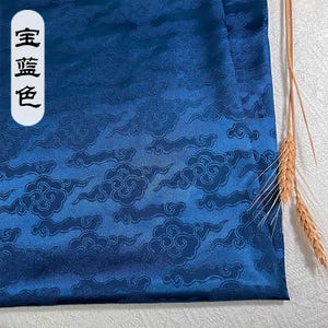 May include: A deep blue satin fabric featuring a repeating tonal jacquard pattern of stylized clouds. A white vertical label on the left side displays the Chinese characters 宝蓝色. A small cluster of dried wheat stalks rests on the right edge.