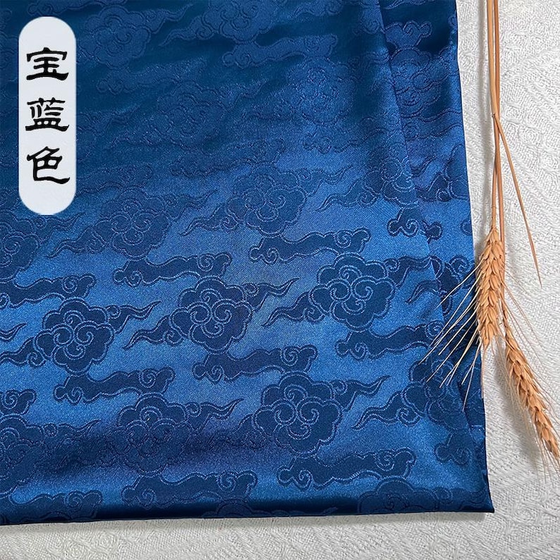 May include: A deep blue satin fabric featuring a repeating tonal jacquard pattern of stylized clouds. A white vertical label on the left side displays the Chinese characters 宝蓝色. A small cluster of dried wheat stalks rests on the right edge.