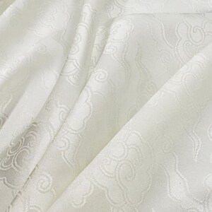 May include: A white fabric featuring a tonal jacquard pattern of stylized clouds. The material has a subtle sheen and is draped to show soft folds, highlighting the intricate woven cloud motifs across the surface of the textile.