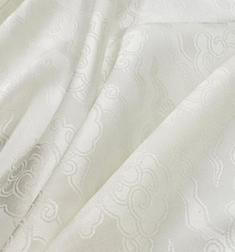 May include: A white fabric featuring a tonal jacquard pattern of stylized clouds. The material has a subtle sheen and is draped to show soft folds, highlighting the intricate woven cloud motifs across the surface of the textile.
