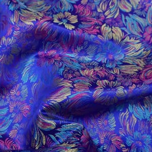 May include: Close-up of a vibrant blue fabric with a floral pattern. The design features stylized flowers in shades of pink, teal, and gold. The fabric has a slight sheen, suggesting a luxurious material suitable for apparel or home decor.