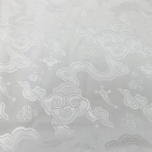 May include: A white fabric featuring a tonal jacquard pattern of stylized swirling clouds and circular coin motifs. The design is monochromatic, with the woven elements creating a subtle texture and sheen against the matte white background surface.