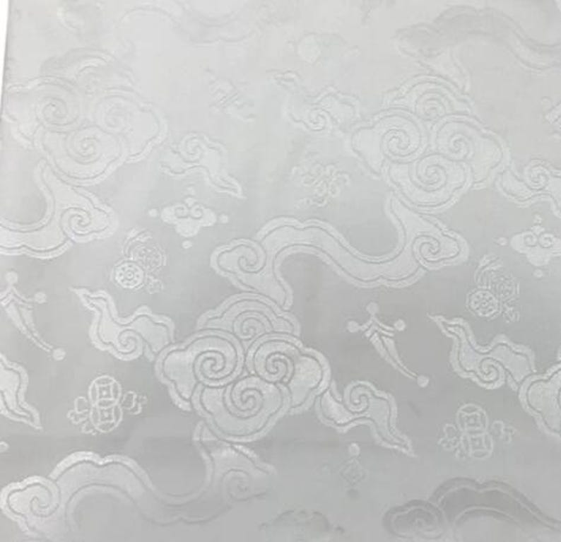 May include: A white fabric featuring a tonal jacquard pattern of stylized swirling clouds and circular coin motifs. The design is monochromatic, with the woven elements creating a subtle texture and sheen against the matte white background surface.