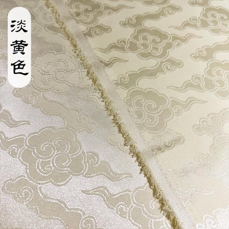 May include: A pale yellow fabric featuring a repeating woven pattern of stylized clouds and swirling wind motifs. The material has a subtle sheen and a frayed edge. A white vertical label on the left side displays the Chinese characters 淡黄色.