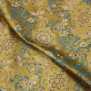 May include: Close-up of a golden fabric with an intricate pattern. The design features stylized birds, flowers, and swirling motifs in shades of teal, white, and gold. The fabric has a subtle sheen.